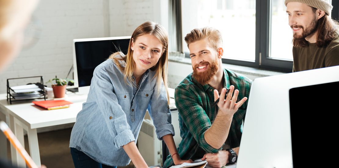 Image of young happy colleagues in office talking with each other using computer. Looking aside.